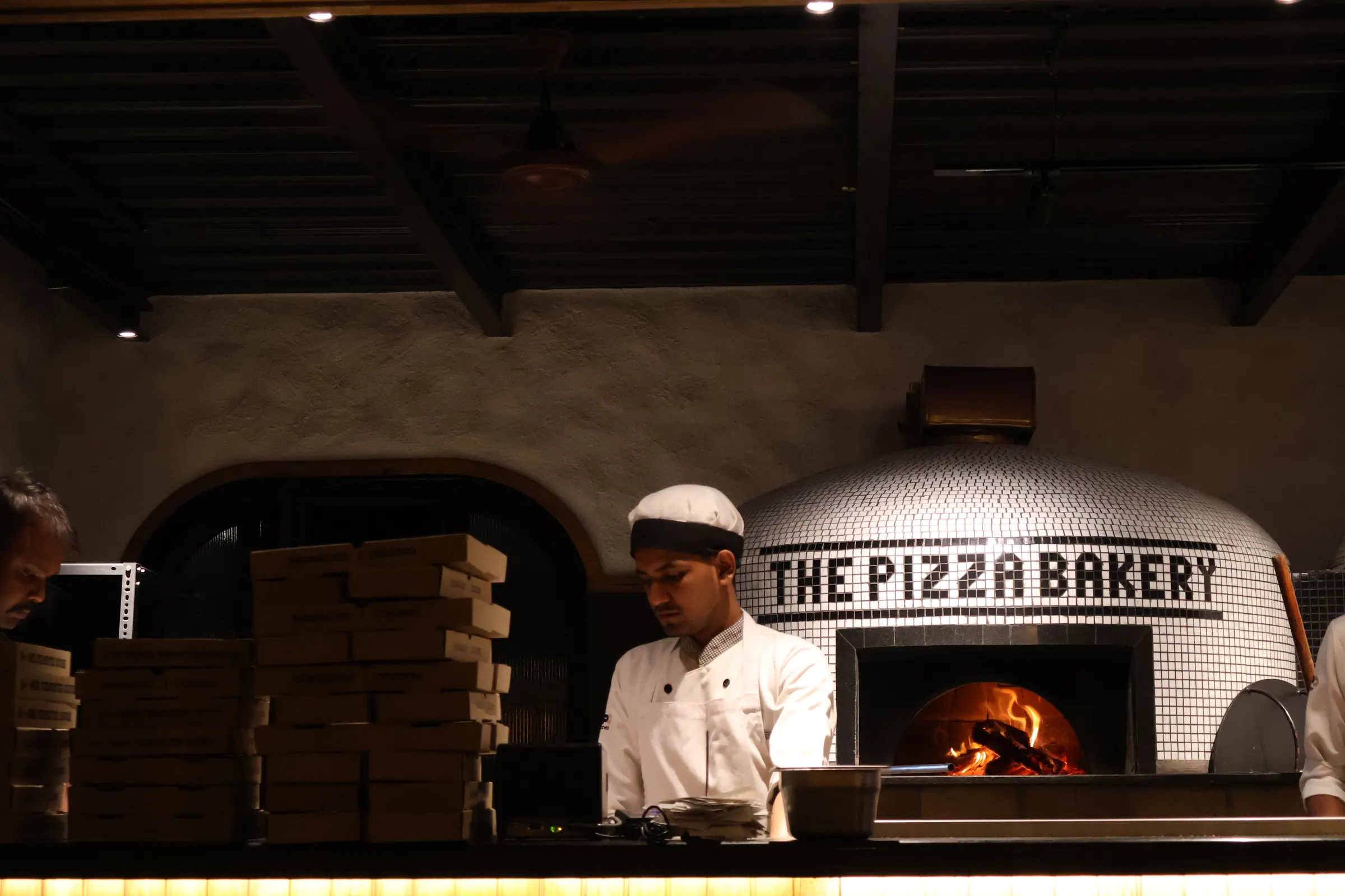 A cook in white kitchen whites and a white cap stands at a tiled wood-fired oven with a fire burning inside it, stacked pizza boxes to his left.