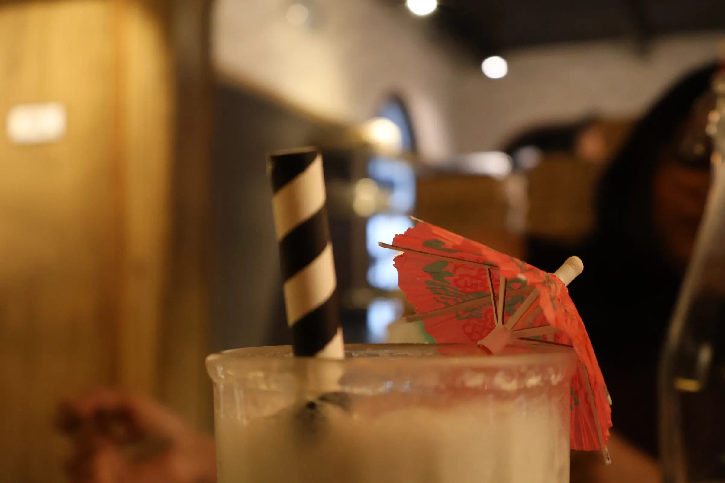 A black and white striped paper straw and a small red paper cocktail umbrella resting on the rim of a frosted glass, with the restaurant behind them thrown out of focus.