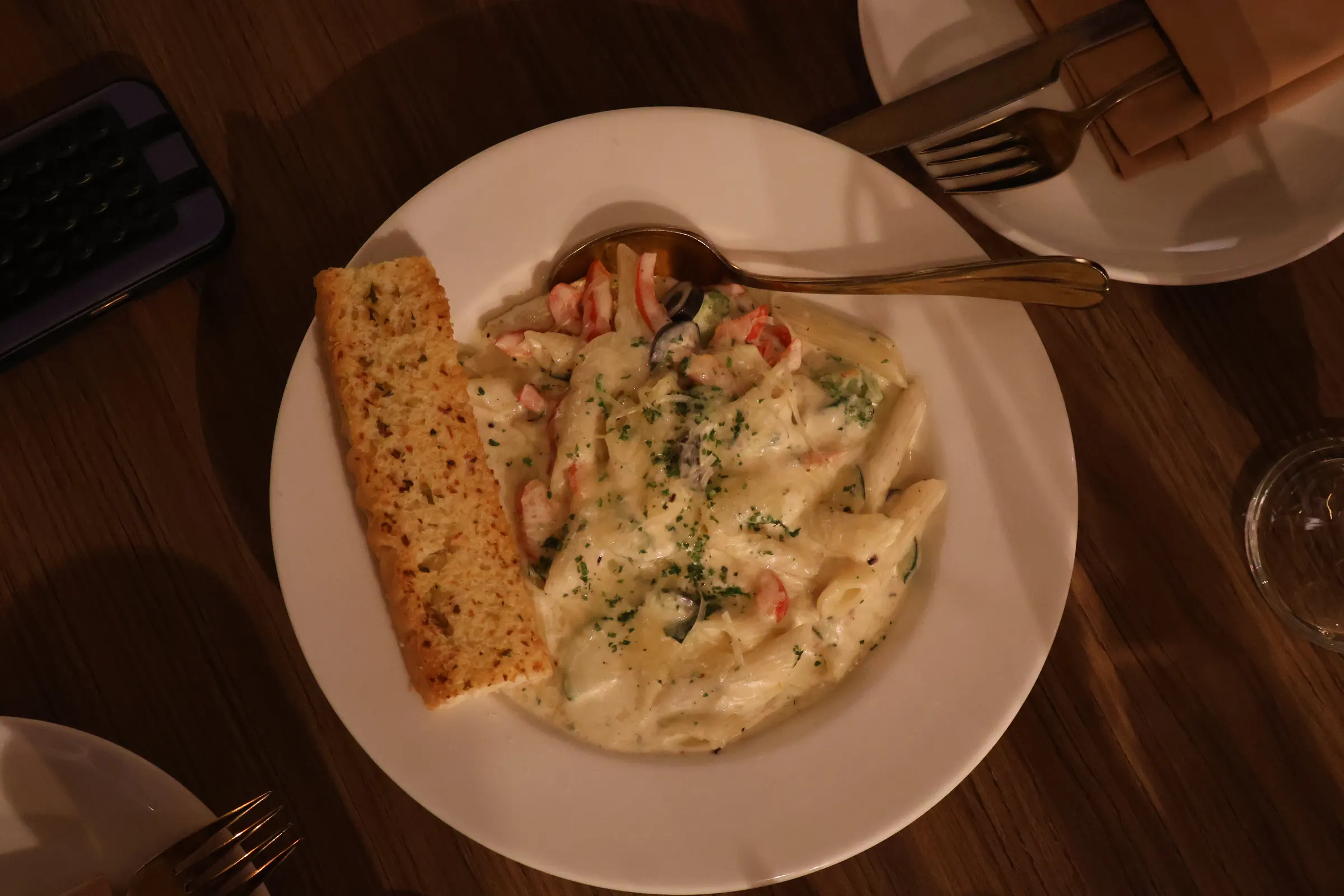 Overhead view of penne in a white sauce with red pepper, olives and courgette, a slice of garlic bread beside it on a white plate, on a dark wood table.