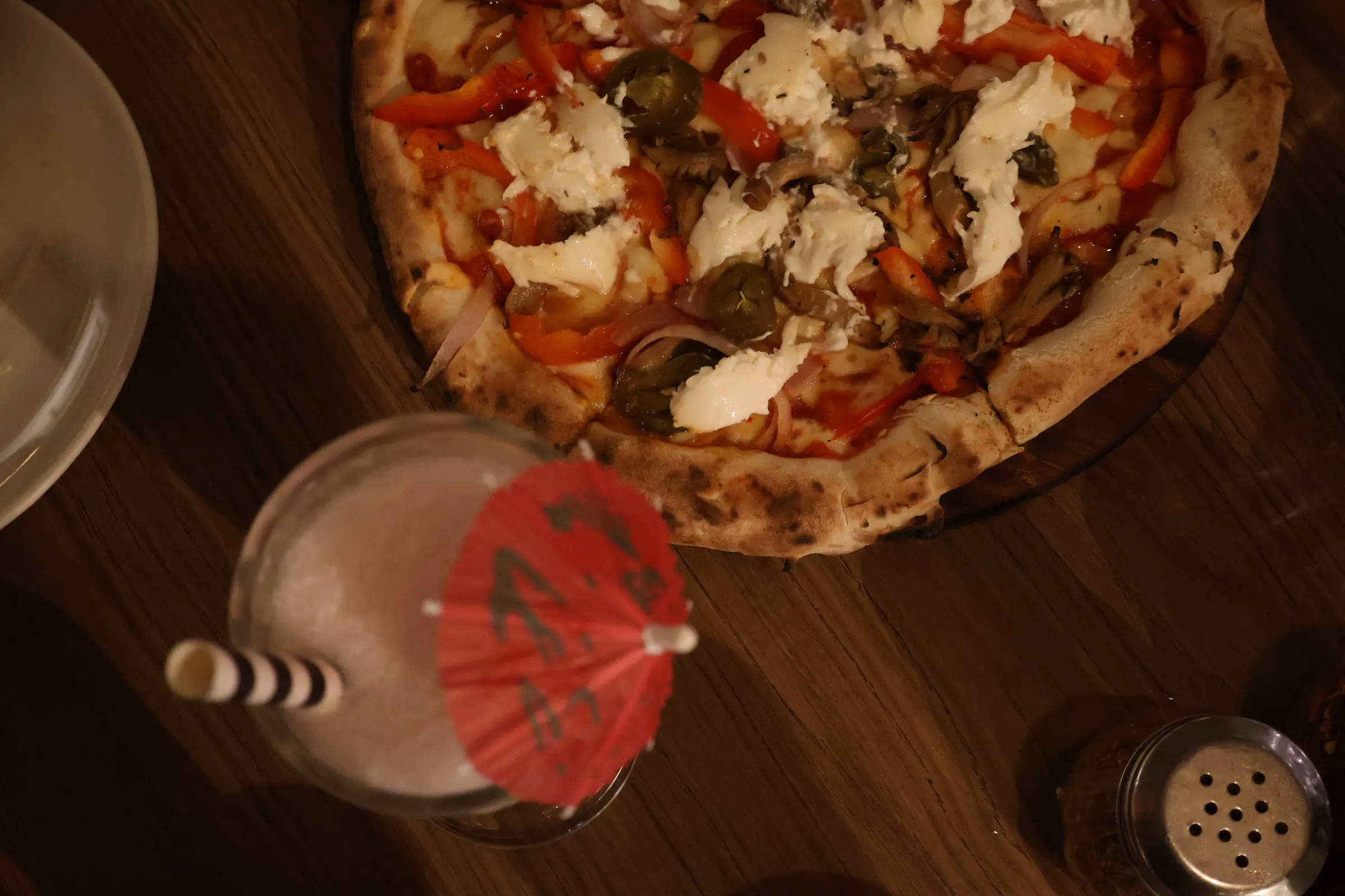 A whole pizza on a wooden board seen from above, topped with red pepper, jalapenos, red onion and torn burrata, with a drink and its paper umbrella at the edge of the frame.